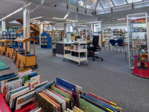 Panorama des Innenraums der Stadtbücherei mit Bücherregalen, einem Schreibtisch und einer großen Holzeisenbahn auf Teppichboden.