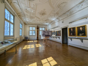 Eleganter Ausstellungsraum im Stadtmuseum mit verzierter Stuckdecke, großen Fenstern, poliertem Holzboden, und Gemälden historischer Persönlichkeiten.