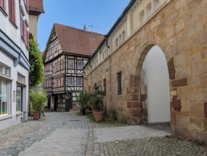 Mittelalterliche Gasse mit unebenem Kopfsteinpflaster, teilweise abgeschliffen für eine ebene Fläche. Der Weg führt zu einem Torbogen rechts. Links daneben ein altes Fachwerkhaus.