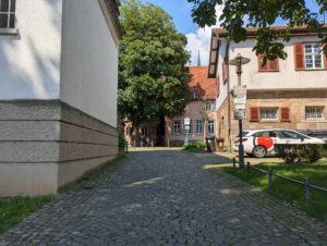 Gehweg mit Kopfsteinpflaster, der zu einem Innenhof führt. Rechts ein Schild mit Pfeil geradeaus und der Aufschrift 'WC'.