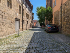 Gehweg mit historischem, unebenem Kopfsteinpflaster, flankiert von alten Sandsteingebäuden. Rechts ein parkendes Auto.