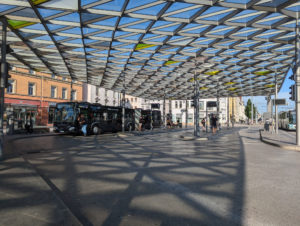 Busbahnhof mit mehreren Haltestellen und zwei parkenden Bussen. Besonders auffällig ist das Dach, das aus vielen kleinen Dreiecken in einer Stahlkonstruktion besteht.